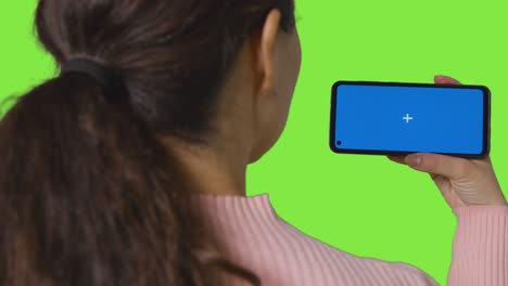 Rear-View-Shot-Of-Woman-Streaming-Content-To-Blue-Screen-Mobile-Phone-Against-Green-Screen